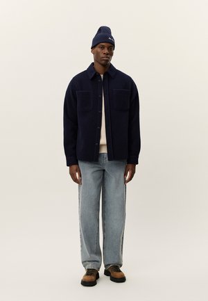 Navy fleece jacket with two front pockets, worn over a light sweater. Paired with light blue jeans and brown lace-up boots, topped with a navy beanie.