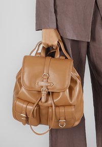 Brun läderryggsäck med en strukturerad design, med en klaffstängning, framfickor och metallhardware i accent. Slät textur, minimal sömnad.