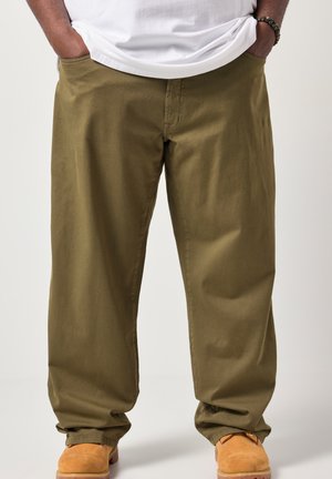 Mężczyzna noszący luźne oliwkowozielone spodnie, beżowe buty robocze, z rękami w kieszeniach i częściowo widoczną białą koszulą.
