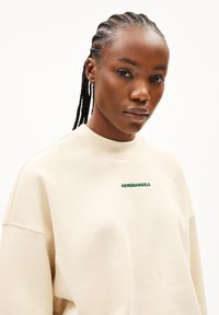 Cremfarbener Sweatshirt mit grünem "ARMEDANGELS"-Text, hochgeschlossen und mit entspannter Passform, aus weichem Baumwollmaterial gefertigt.