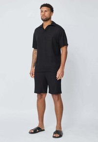 Polo shirt noir en lin à manches courtes associé à un short noir assorti et des sandales. Design minimaliste, coupe décontractée et texture douce.
