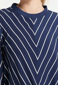 Marineblaues Oberteil mit weißen vertikalen und diagonalen Streifen im Chevron-Muster, mit einem runden Ausschnitt und weichem, strukturiertem Stoff.