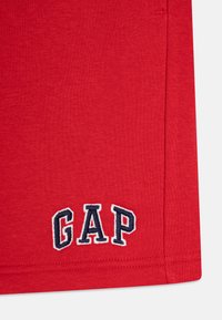 Rød bomull T-skjorte med en marineblå brodert "GAP"-logo nederst til venstre. Glatt tekstur, korte ermer og klassisk passform.