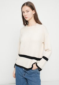 Beżowy dzianinowy sweter z czarnymi poziomymi paskami, okrągłym dekoltem i opuszczonymi ramionami. Miękka tekstura i zaokrąglony dół swetra na niebieskich dżinsach.