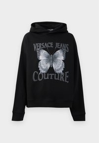 Melna kapuce, izgatavota no mīksta auduma, ar lielu pelēku tauriņa grafiku un „VERSACE JEANS COUTURE” tekstu treknā fontā.