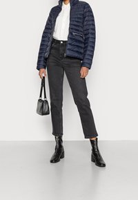 Giacca puffer navy con cuciture orizzontali, due cerniere laterali, camicia bianca, jeans neri a vita alta, stivali neri con tacco e una piccola borsa.