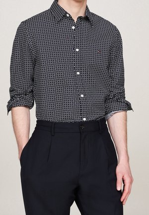 Mężczyzna w czarnej koszuli na guziki z długim rękawem z białymi owalnymi kropkami i czarnych spodniach z wysokim stanem, ręce w kieszeniach.