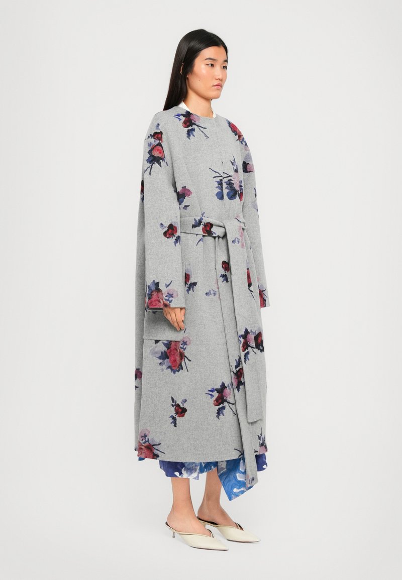 Donna che indossa un lungo cappotto grigio a fiori con una cintura in vita, in piedi di lato, abbinato a una gonna blu a fiori e scarpe con tacco kitten bianche a punta.