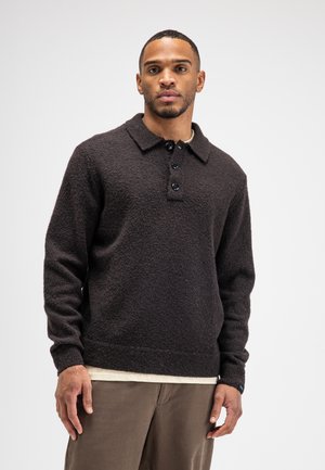 Man wearing a dark textured long-sleeve sweater with a collar and three buttons, beige undershirt, and brown pants against a white background.