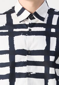 Chemise boutonnée avec un fond blanc présentant un motif à carreaux bleu marine audacieux. Elle a un col pointu et des boutons noirs.