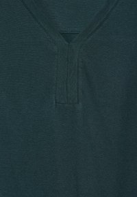 Teeppari, jossa on tehosteväri turkoosi, avara V-pääntie ja näkyvät saumat. Pehmeä rakenne, lyhyet hihat ja siisti viimeistely pääntiellä.