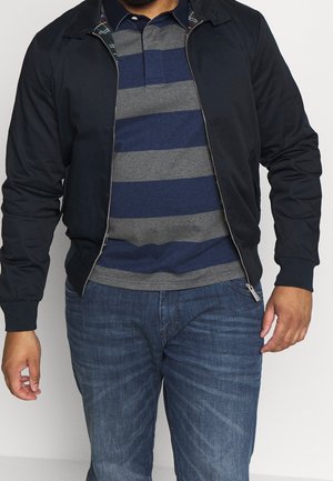 Man wearing navy blue and gray striped polo shirt, dark navy zip-up jacket, and blue jeans against a plain background.