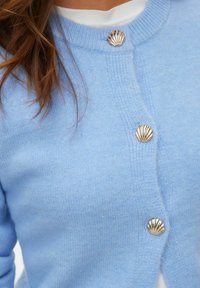 Cardigan en maille bleu clair avec un col rond et trois boutons dorés en forme de coquillage. Texture douce, coupe ajustée et poignets côtelés.