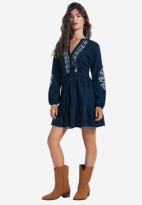 Abito in lino blu navy con maniche lunghe, dettagli ricamati, cintura in vita e gonna svasata. Abbinato a stivali da cowboy in suede marrone.