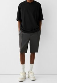 T-shirt noir surdimensionné associé à un short gris mi-cuisse. Le modèle porte des baskets blanches avec des accents clairs et des chaussettes blanches côtelées.