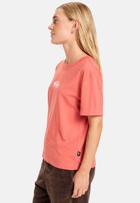 Koraalroze T-shirt met korte mouwen van katoen, met een ronde Hals en een klein wit logo op de borst. Glad textuur en een comfortabele pasvorm.