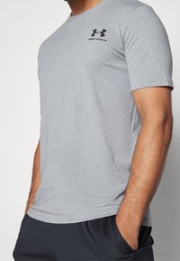 Hombre vistiendo una camiseta ajustada de color gris claro de Under Armour con un logo negro, con las manos en los bolsillos, cortada en el cuello y la cintura.