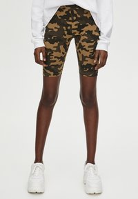 Brune og sorte camouflagemønstrede cykelshorts lavet af elastisk stof, med en mellemhøj talje og en tætsiddende pasform, kombineret med hvide sneakers.