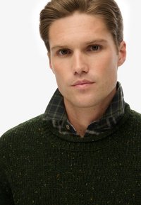 Pull en laine vert avec des éclats multicolores, présentant un col haut et une chemise à carreaux en dessous, principalement en vert foncé et gris.