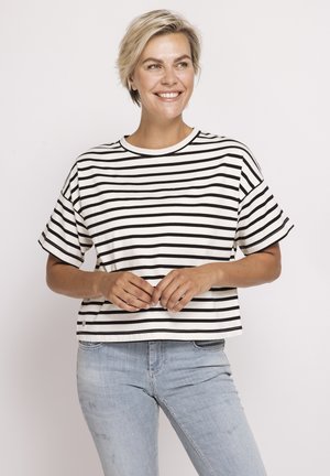 Frau mit kurzen blonden Haaren, die ein schwarz-weiß gestreiftes Kurzarmshirt und hellblaue Jeans trägt, lächelt und hält die Hände zusammen.