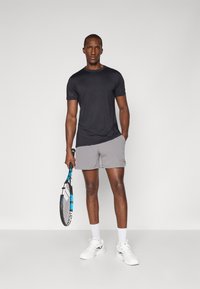 Schwarzes, kurzärmeliges Sportshirt, graue Shorts, hält einen Tennisschläger. Die Schuhe sind weiß mit hellblauen Akzenten am Schlägergriff.