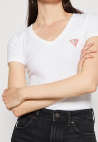 Camiseta blanca de manga corta con un escote en V profundo y un logo triangular rojo en el pecho, combinada con jeans de mezclilla oscura.