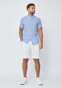 Chemise à rayures bleues et blanches à manches courtes avec un col montant, accompagnée de shorts blancs. Le modèle porte des baskets blanches, se tenant devant un fond clair.