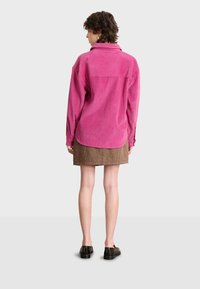 Camisa de pana rosa de manga larga con cuello, combinada con una falda de cuadros marrón. Textura suave y detalles acanalados. Zapatos negros.