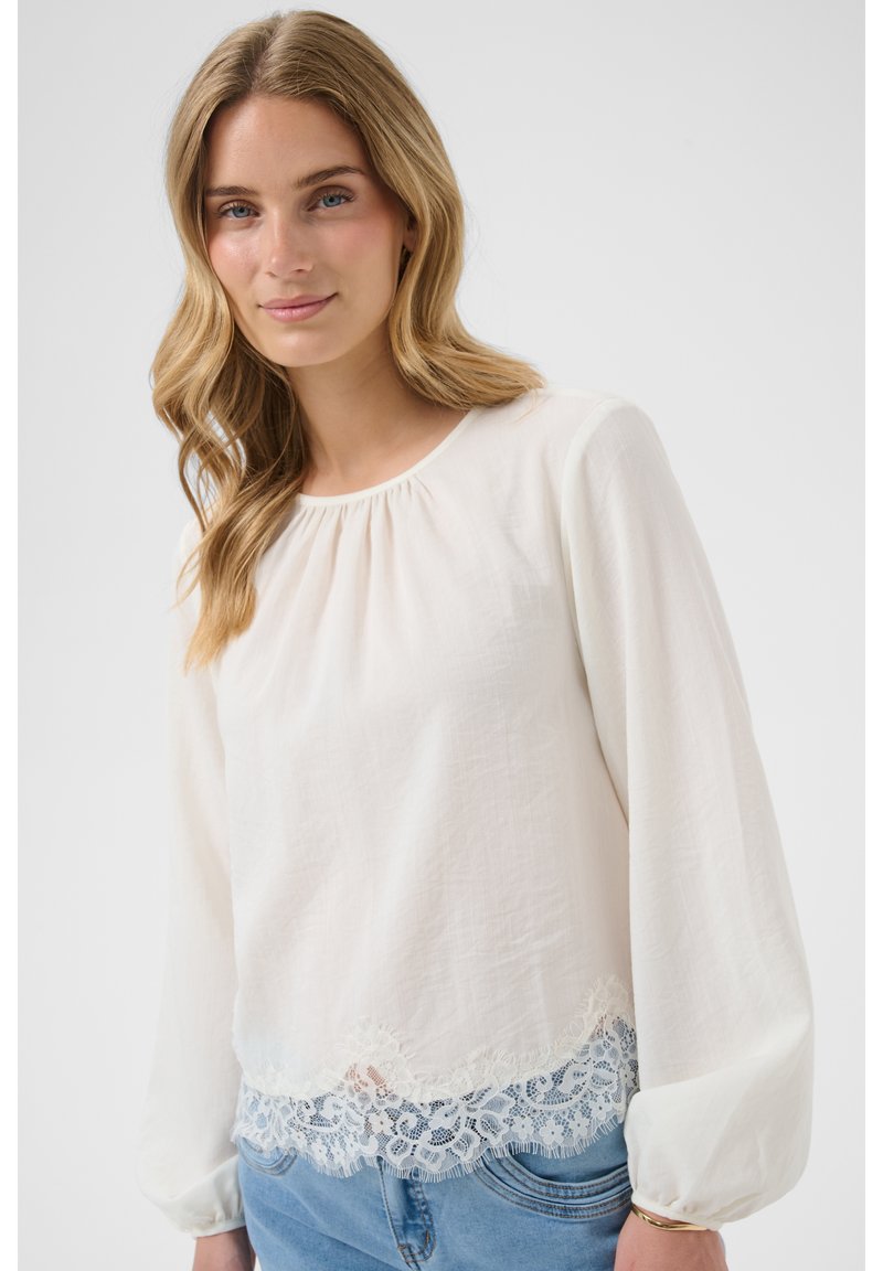 Jeune femme aux longs cheveux blonds porte un chemisier blanc avec dentelle et un jean bleu clair sur un fond uni.