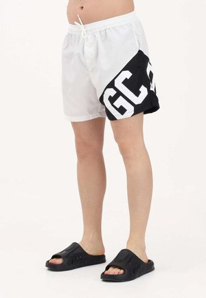 Person trägt weiße und schwarze Shorts mit großen "GC" Buchstaben an der Seite, kombiniert mit schwarzen offenen Sandalen zum Hereinschlüpfen.