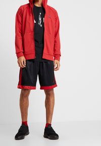 Sudadera roja con cremallera y capucha, camiseta negra con un gráfico, pantalones cortos negros con detalles rojos y zapatillas deportivas negras con toques rojos.