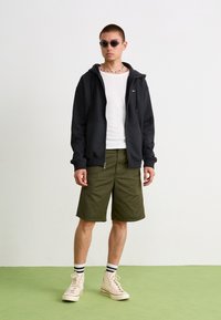 Sweatshirt preto com fecho, camisa branca, calções verde-oliva, ténis bege com meias às riscas pretas e brancas, óculos de sol redondos, colar de contas.