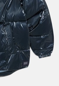 Marineblå pufferjakke med en skinnende finish, quiltet design, elastiske manchetter og sidelomme. Brandetiket vist på venstre side.