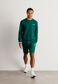 Grøn sweatshirt og shorts sæt i bomuld med hvide detaljer, der har en rund halsudskæring og logodetalje. Modellen går foran en neutral baggrund.