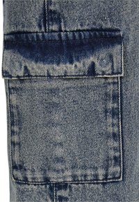 Denim fabric with a light blue wash, featuring a patch pocket on the thigh, reinforced stitching, and a slightly faded texture.