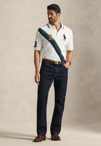 Άνδρας που φοράει λευκό πόλο με πράσινες και ναυτικό μπλε διαγώνιες ρίγες, σκούρα τζιν, καφέ ζώνη, καφέ μοκασίνια και ρολόι καρπού, στέκεται με το ένα χέρι στην τσέπη.