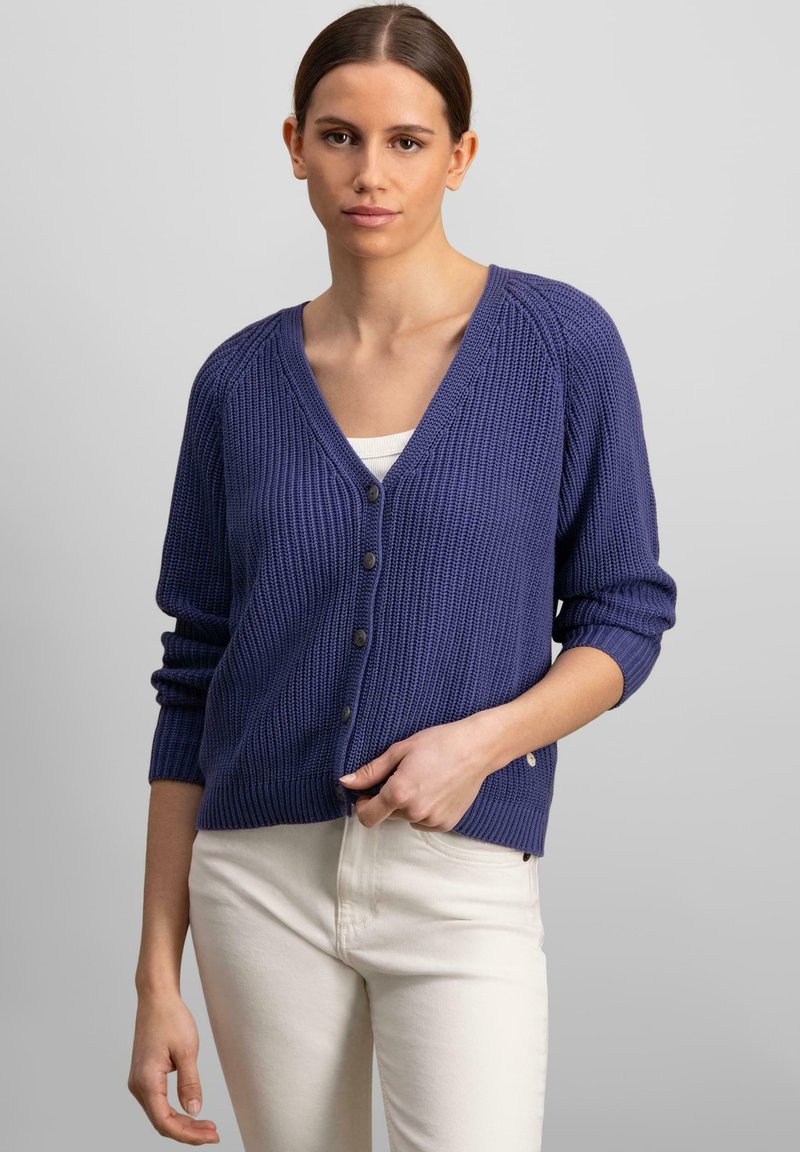 Fialový pletený cardigan s výstrihom do V, štyrmi gombíkmi na prednej strane a ryhovanou textúrou. V kombinácii so svetlými nohavicami.