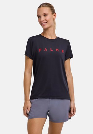 Femme aux cheveux blonds attachés en queue de cheval, portant un t-shirt noir Falke et un short de sport gris, souriante et regardant sur le côté.