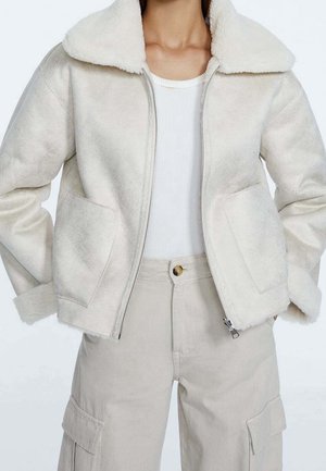 Femme portant une veste beige zippée avec de grandes poches sur un top blanc côtelé, associée à un pantalon cargo beige.