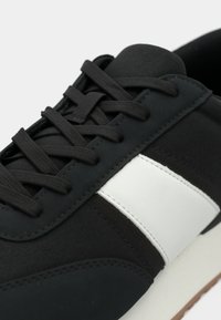 Μαύρο αθλητικό παπούτσι με λευκή ρίγα, με υφή υφάσματος, κορδόνια και λάστιχο σόλα. Διακριτική ραφή στο επάνω μέρος.