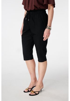 Vrouw draagt zwarte knielange broek met trekkoord, bruin topje, zwarte sandalen met hak en gouden accenten, en meerdere armbanden aan haar rechterpols.
