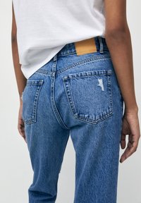 Jeans de denim azul claro con un corte relajado, que cuentan con un parche de cuero marrón, bolsillos traseros y sutiles detalles desgastados.