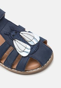 Sandalias azul marino con diseño de punta abierta, hechas de material suave. Presentan un bug bordado decorativo en azul y plata en la parte delantera.