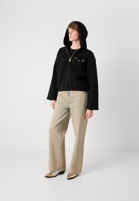 Pulôver preto com capuz, feito de tecido texturizado e com um fecho dourado. Usado com jeans de perna larga em azul claro e sapatos brilhantes de bico fino.