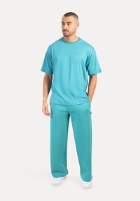 Man som står och bär en matchande överdimensionerad t-shirt i teal och vida byxor med vita sneakers, ser något åt sidan.