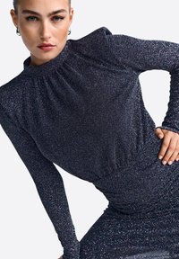 Vestido negro brillante con mangas largas, cuello alto y detalles fruncidos en los hombros y la cintura. Fabricado con una tela texturizada brillante.