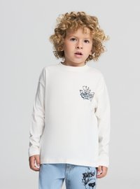 Biała bawełniana koszula z długim rękawem i graficznym nadrukiem na lewej piersi. Do tego jasnoniebieskie jeansy z nadrukowanym wzorem.