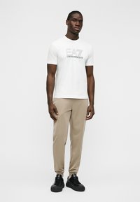 Λευκό κοντομάνικο t-shirt με μαύρο λογότυπο EA7, συνδυασμένο με μπεζ παντελόνια με στενή γραμμή και μαύρα αθλητικά παπούτσια, σε απλό φόντο.