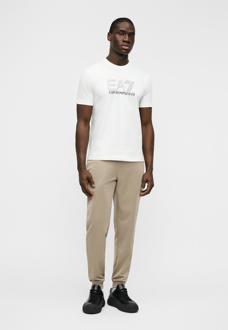 Λευκό κοντομάνικο t-shirt με μαύρο λογότυπο EA7, συνδυασμένο με μπεζ παντελόνια με στενή γραμμή και μαύρα αθλητικά παπούτσια, σε απλό φόντο.