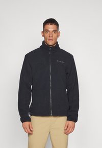 Veste en polaire noire avec un col montant et une fermeture éclair à l'avant. Elle possède des manches longues et un logo sur le côté gauche de la poitrine. Tissu doux et texturé.